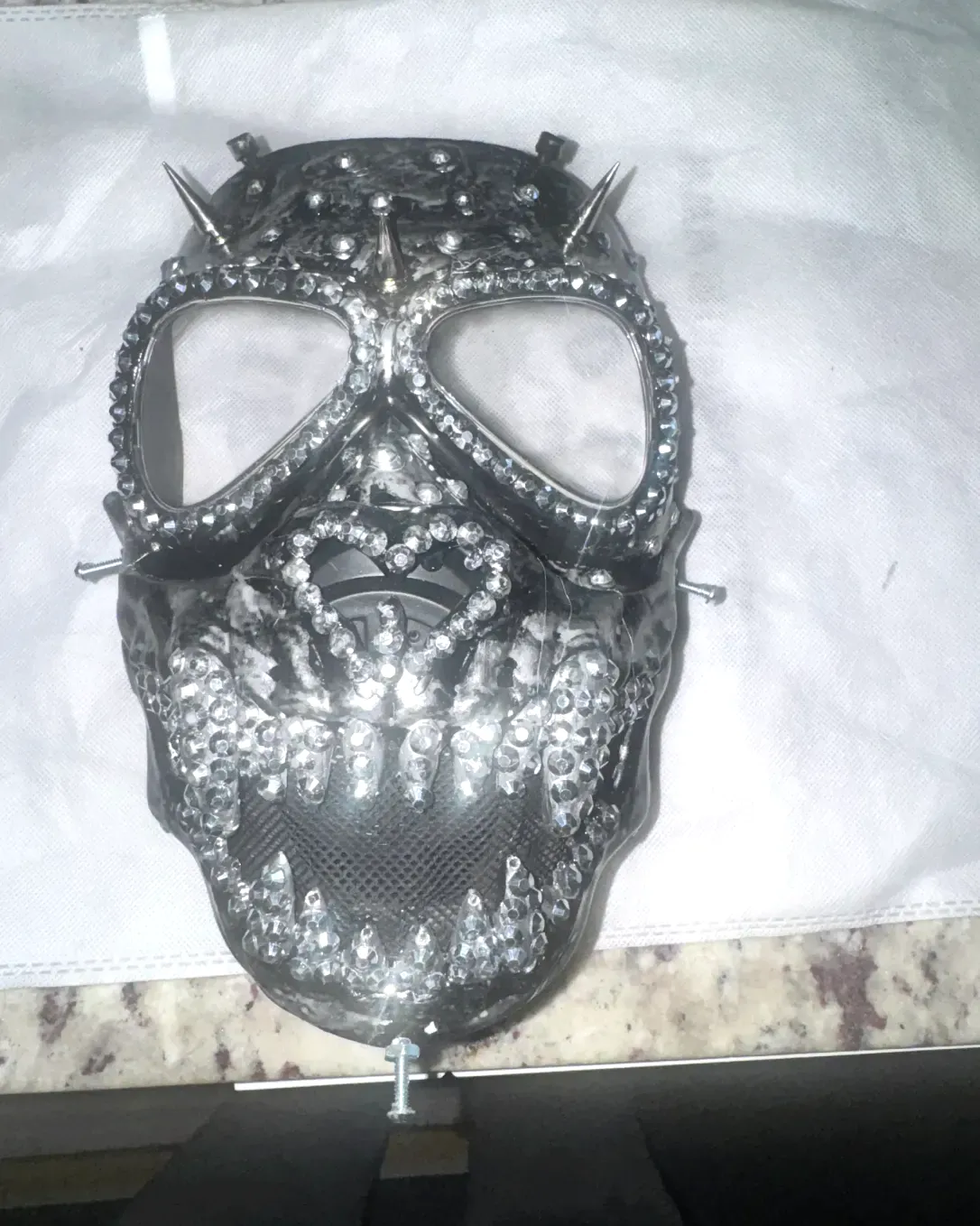 Flat-lay view of the black and gunmetal gas-style mask with crystal-lined eye rims, skull mouth, and crown spikes