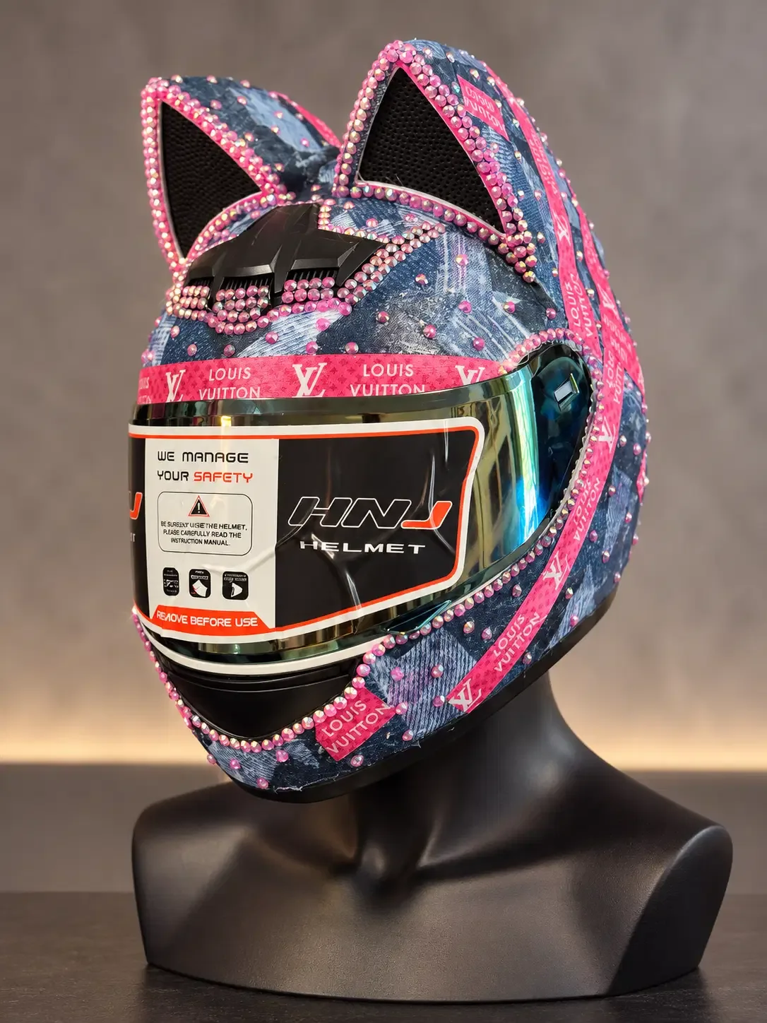 Three-quarter view of a denim-finish cat-ear helmet with pink crystal trim and an iridescent shield on a black mannequin bust