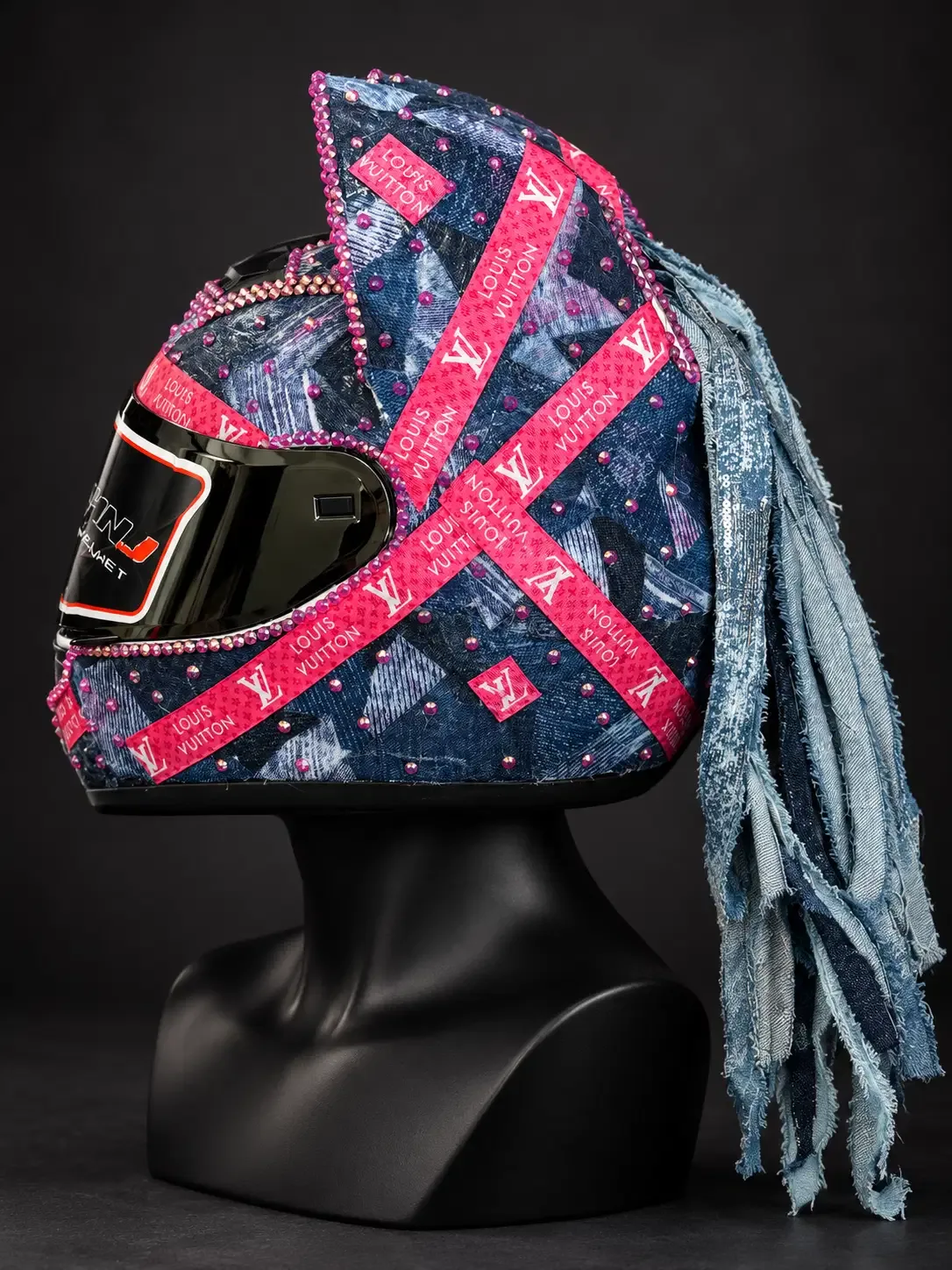 Side view of a denim-finish cat-ear helmet with pink crystal trim and a long frayed denim fringe mohawk on a black mannequin bust