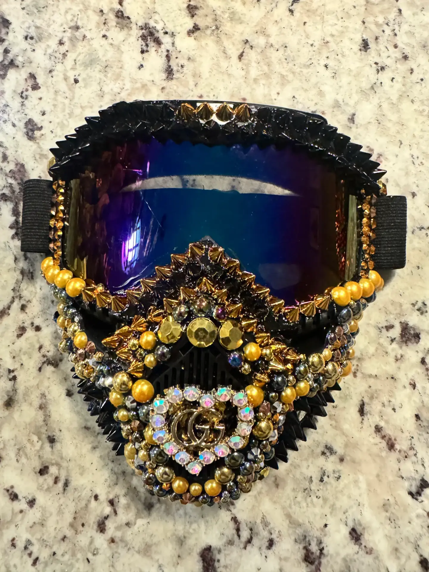 Close-up detail of a black and gold specialty mask showing its dark iridescent lens, layered metallic beads, crystal accents, and sharp spikes
