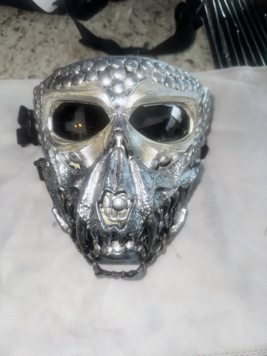 Flat-lay view of the gunmetal two-face skull mask showing its layered metal plating and dark lenses