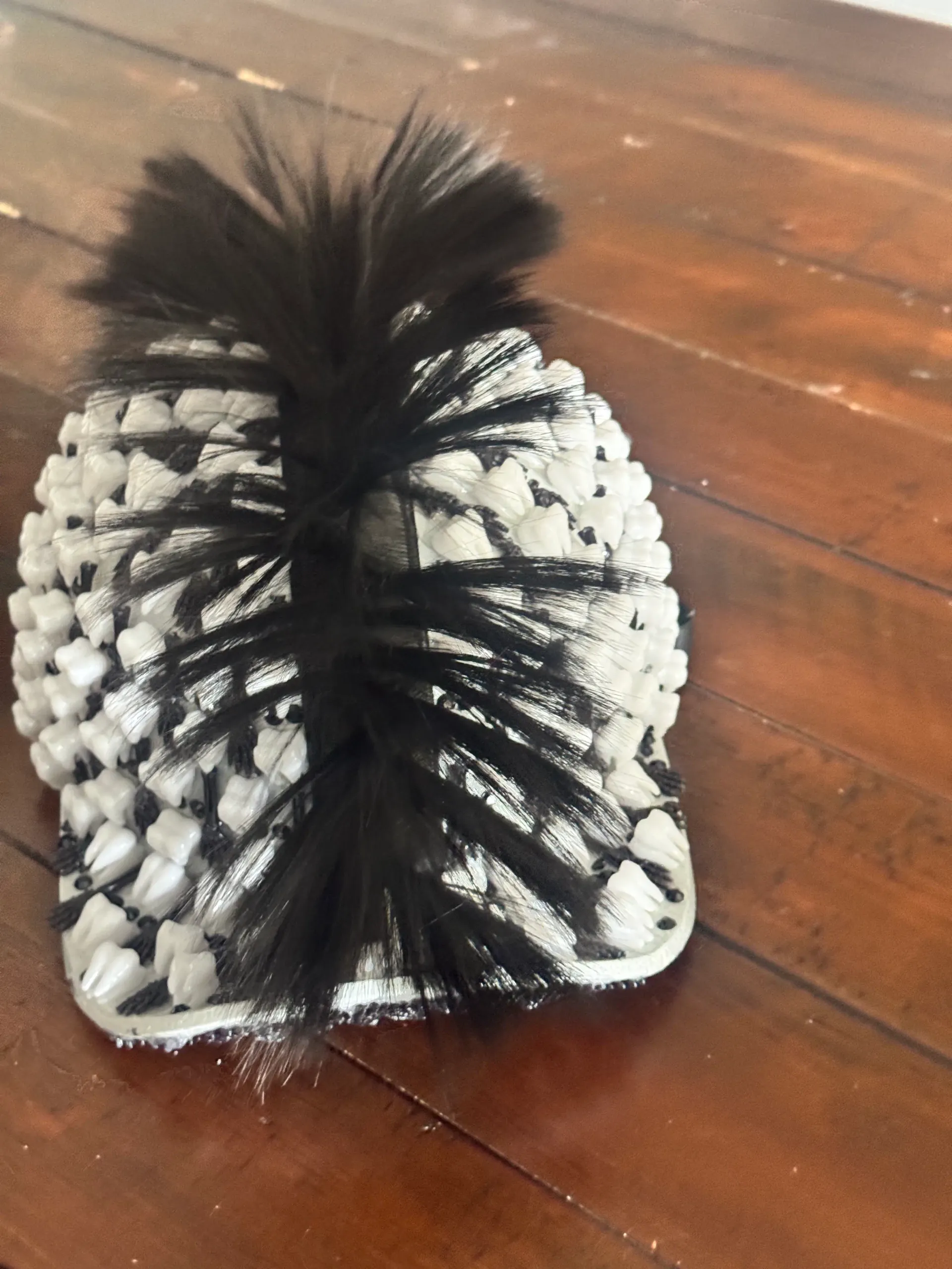 Back detail of the black teeth helmet shell with layered white teeth, black bead trim, and feather mohawk on a wood table